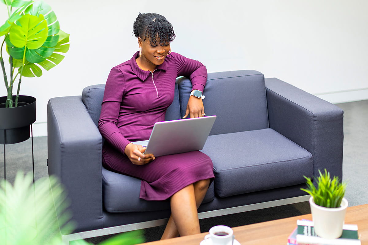 A student in a purple dress sits on a gray sofa with a laptop, smiling. The room is bright with green plants, conveying a relaxed, productive atmosphere.