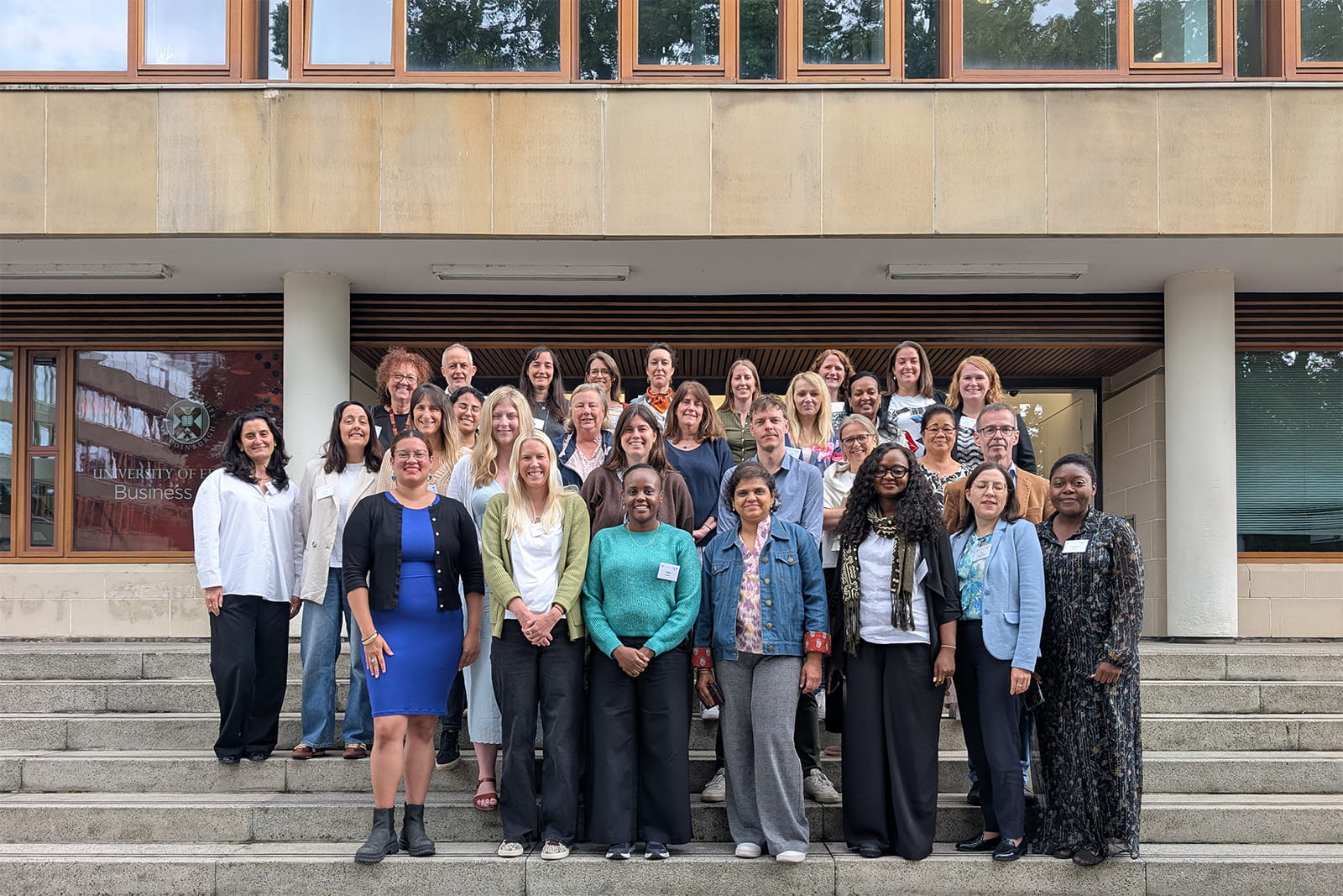 A large group of conference delegates standing on stone steps outside the University of Edinburgh Business School, smiling towards the camera. The Business School name and University crest are visible on the glass windows behind them.
