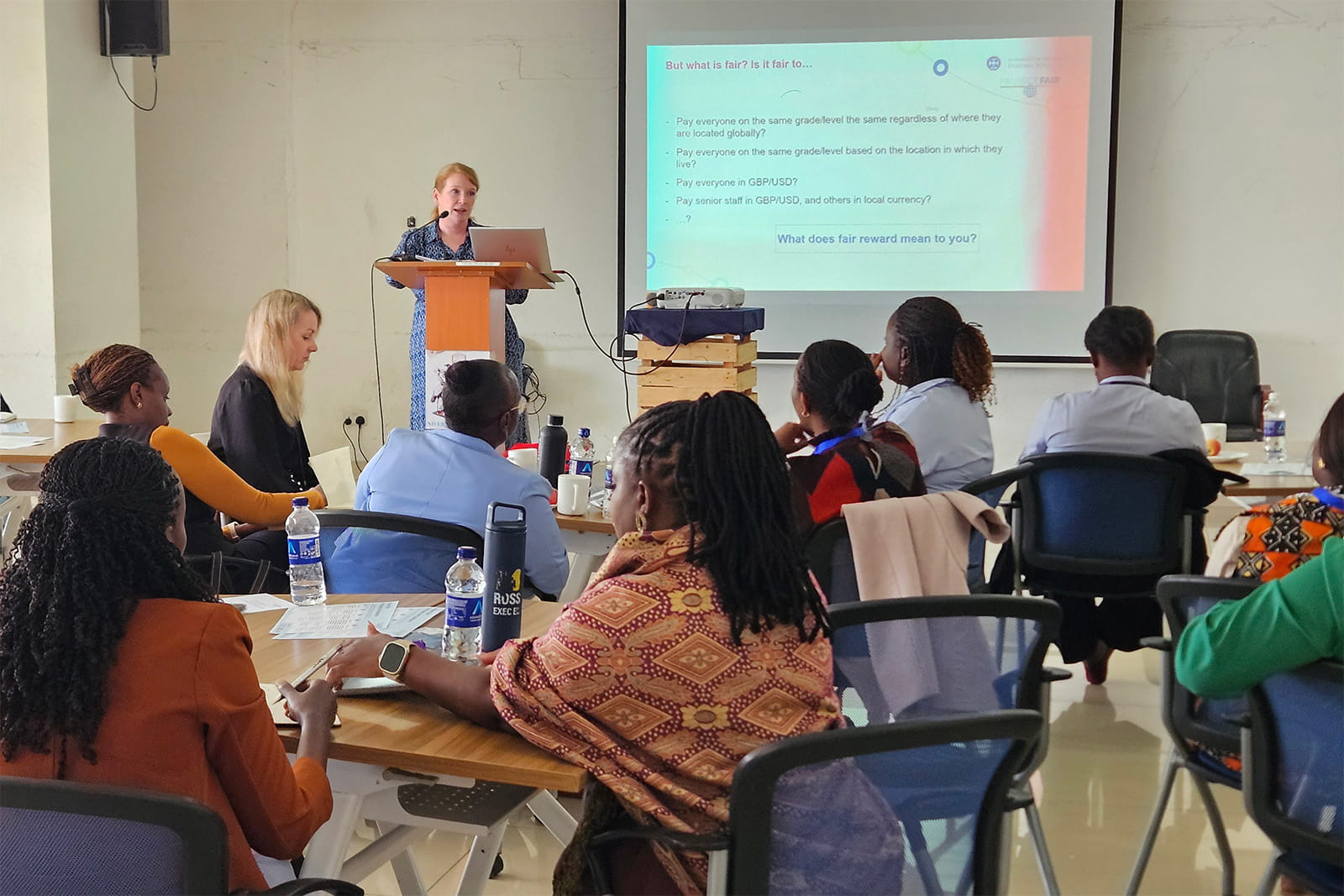 Ishbel stands at a wooden lectern with a laptop, speaking to participants seated around tables in a workshop room. A projected slide behind her reads 'But what is fair? Is it fair to…' and lists questions about paying staff on the same grade globally versus by location, and in GBP/USD versus local currency, ending with 'What does fair reward mean to you?'