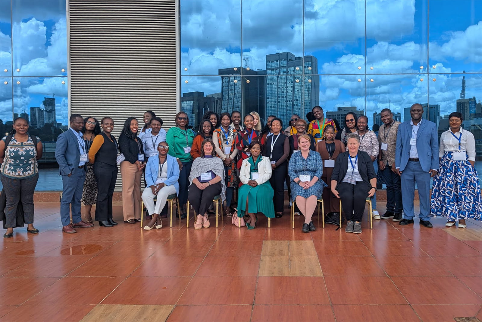 A large group of around 25 workshop delegates posing together on an outdoor terrace with a red-tiled floor. Behind them, a glass wall reflects the Nairobi city skyline, including several high-rise towers, under a blue sky with white clouds. Five people are seated on chairs at the front, with the rest standing.